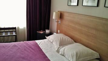 Una camera d'albergo con un letto, un comodino, una lampada, una sedia e una finestra con tende.