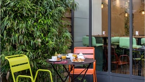 Una mesa con alimentos para el desayuno, dos sillas y una planta de bambú grande al fondo.