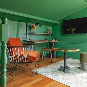A room with a green wall, a wooden floor, a round table with a metal base, a chair with a cushion, a television mounted on the wall, and a shelf with decorative items.