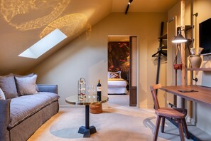 A cozy room with a sofa, a small round table with a bottle and glasses, a wooden chair, and a bed with a patterned headboard.