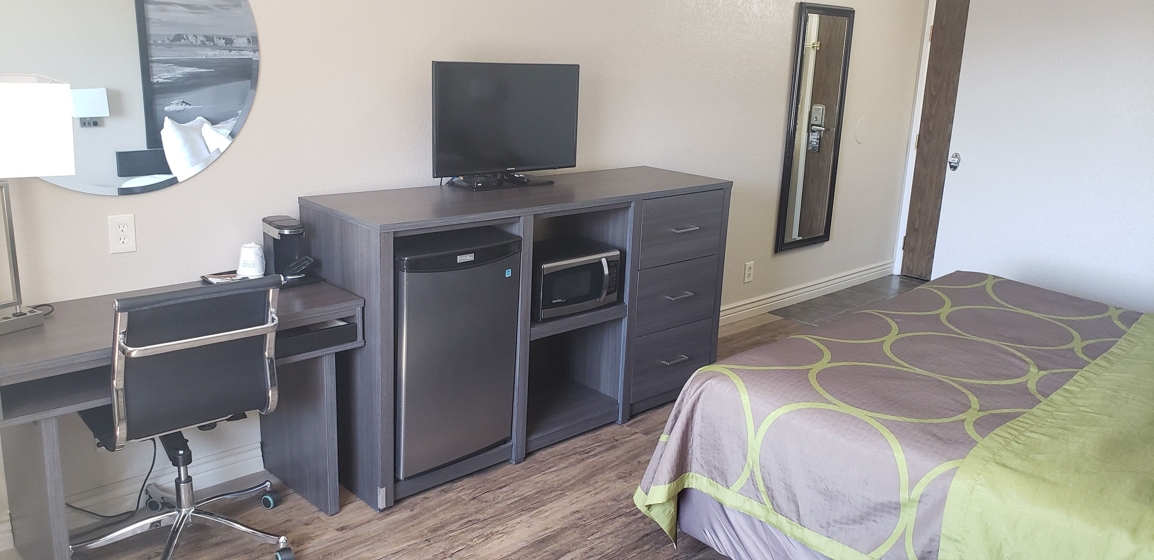 A hotel room with a bed, a desk with a computer, a refrigerator, a microwave, and a television.