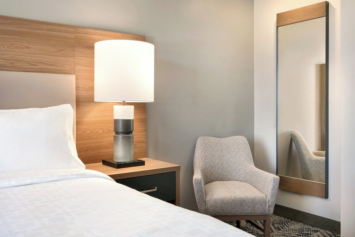Une chambre d’hôtel avec un lit, une table de chevet avec une lampe, une chaise et un grand miroir.