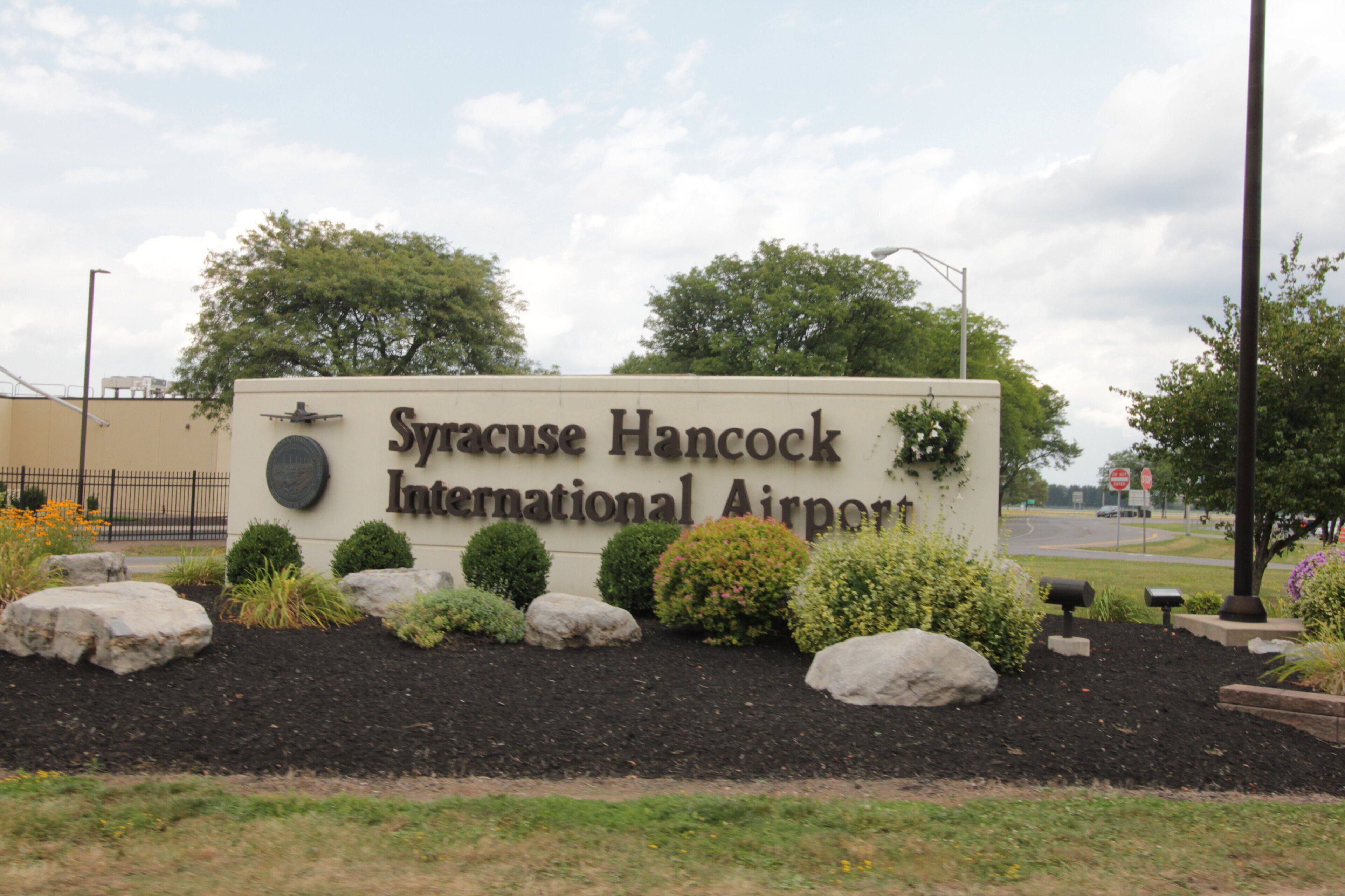 A sign for Syracuse Hancock International Airport surrounded by landscaped areas with rocks and shrubs.