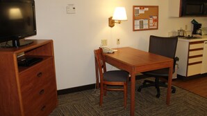 Une chambre d’hôtel avec un bureau en bois, une chaise, un téléviseur, une lampe, un tableau d’affichage et une kitchenette.