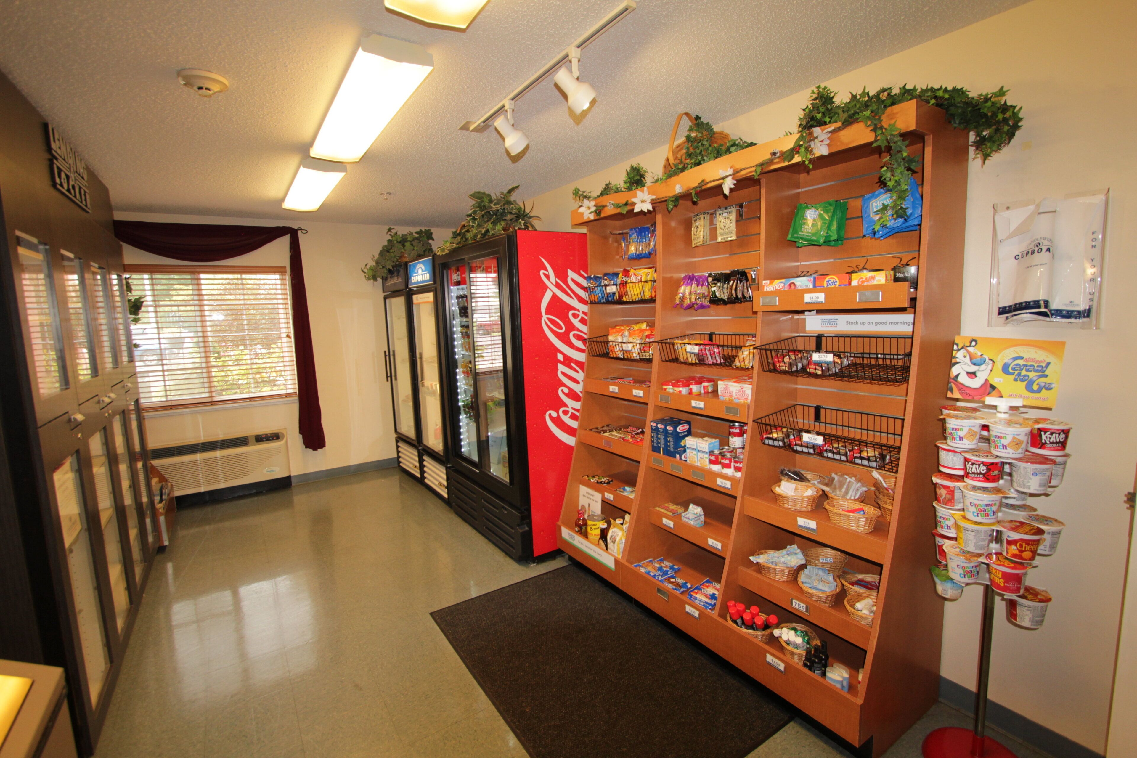 A grocery store aisle with shelves stocked with snacks and beverages, a Coca-Cola refrigerator, and a vending machine.