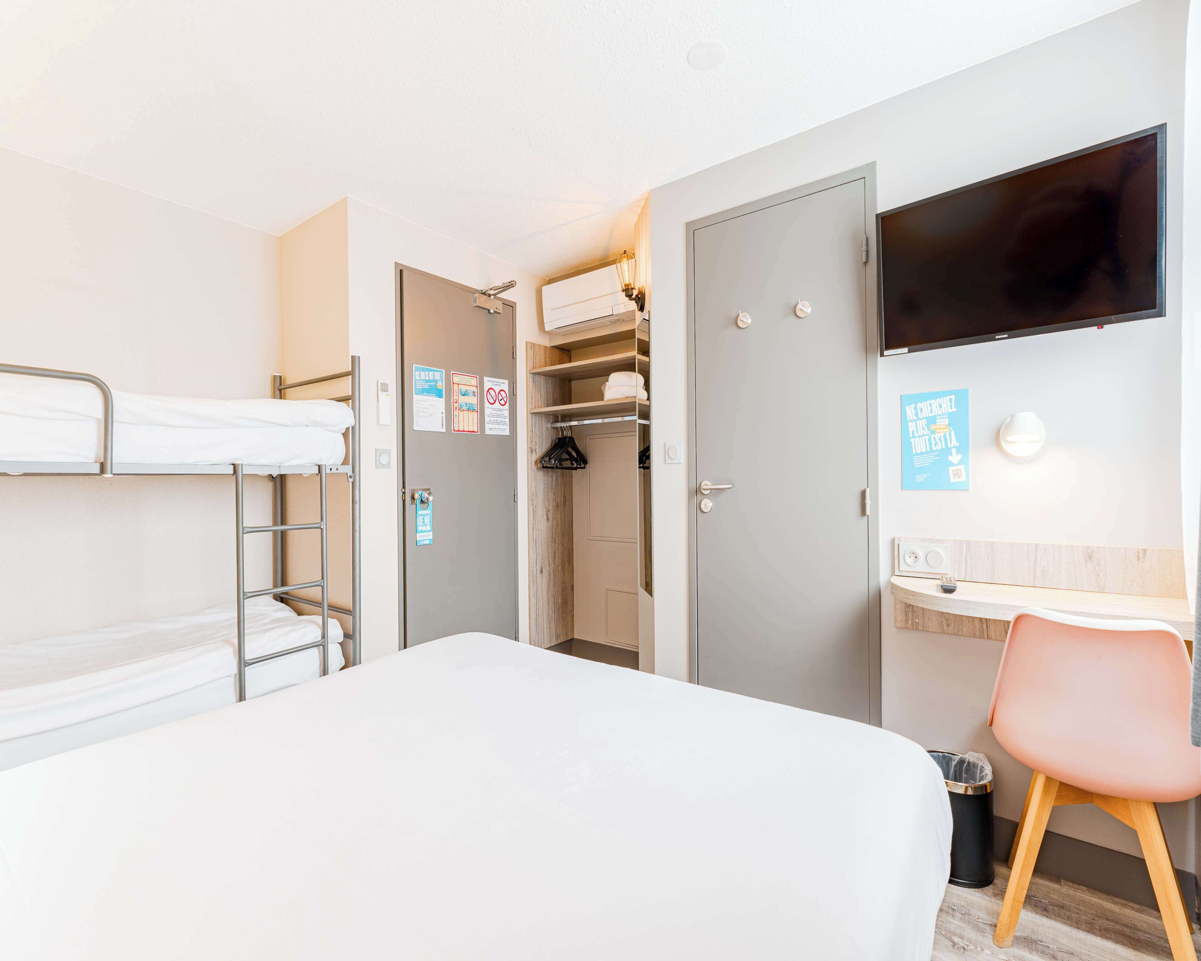 A hotel room with a bunk bed, a desk with a chair, a TV mounted on the wall, and a closet with a mirror.