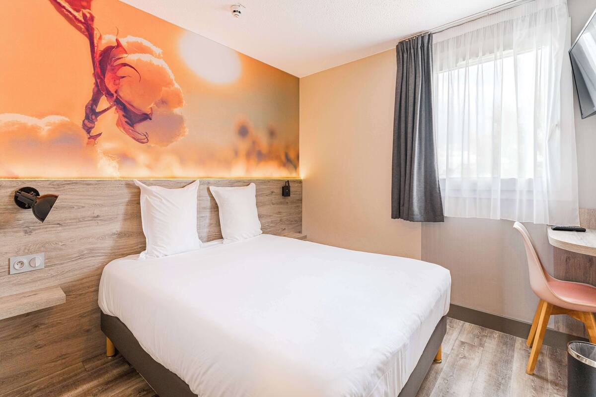 A hotel room with a bed, a wall-mounted lamp, a desk with a chair, and a large window with curtains.