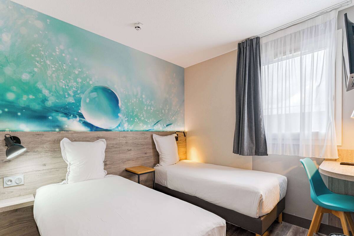 A hotel room with two beds, a wooden headboard, a wall mural, a window with curtains, a desk with a chair, and a trash bin.