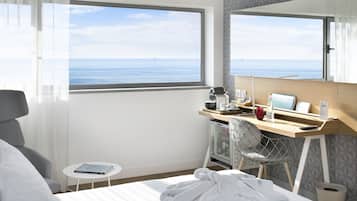 A hotel room with a bed, a desk with a computer, a chair, a small table, and a view of the sea.