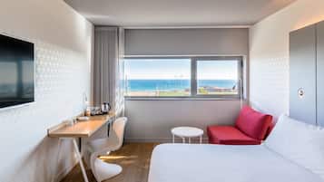 A hotel room with a bed, a desk with a chair, a small table, and a view of the sea.