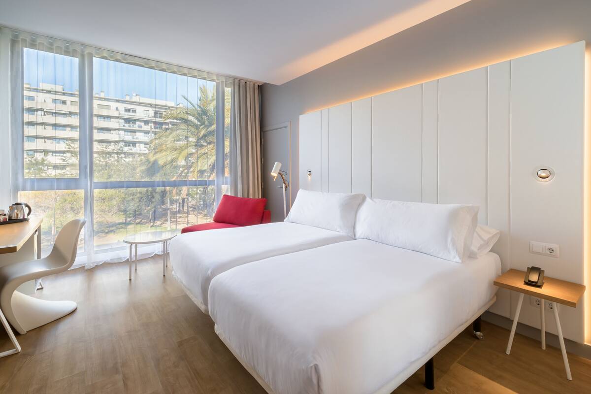 A hotel room with two beds, a desk, a chair, a small table, and a window with a view of buildings and trees.