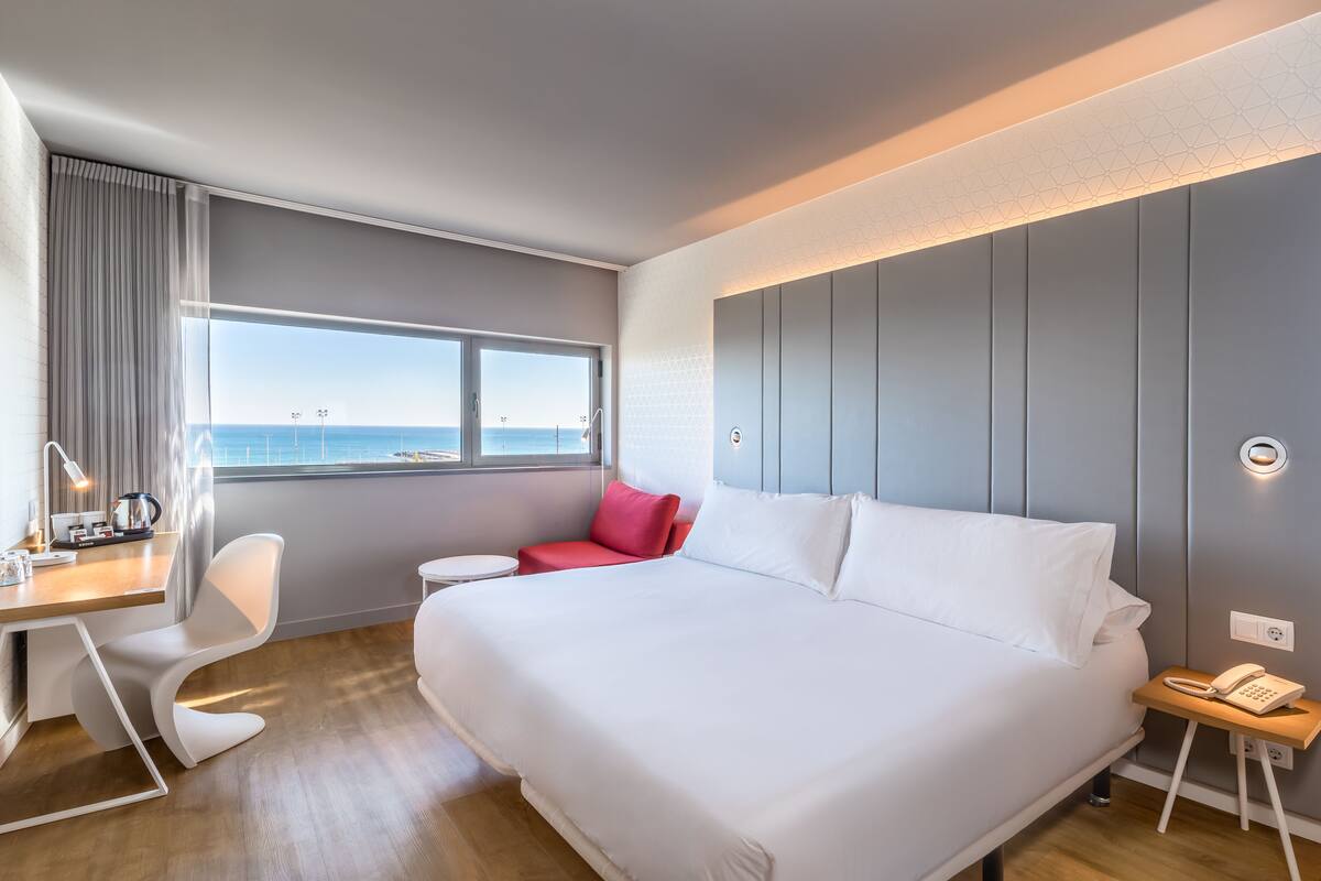 A modern hotel room with a large bed, a desk with a chair, a small red sofa, and a view of the sea through a large window.
