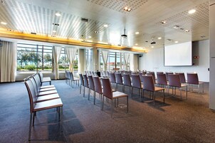 Un salón de conferencias con filas de sillas, una pantalla de proyector y ventanas amplias.