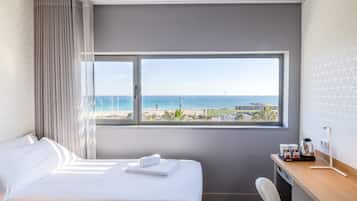 Una habitación de hotel con cama, escritorio, silla, ventana con vista a la playa y al océano, y cortina.
