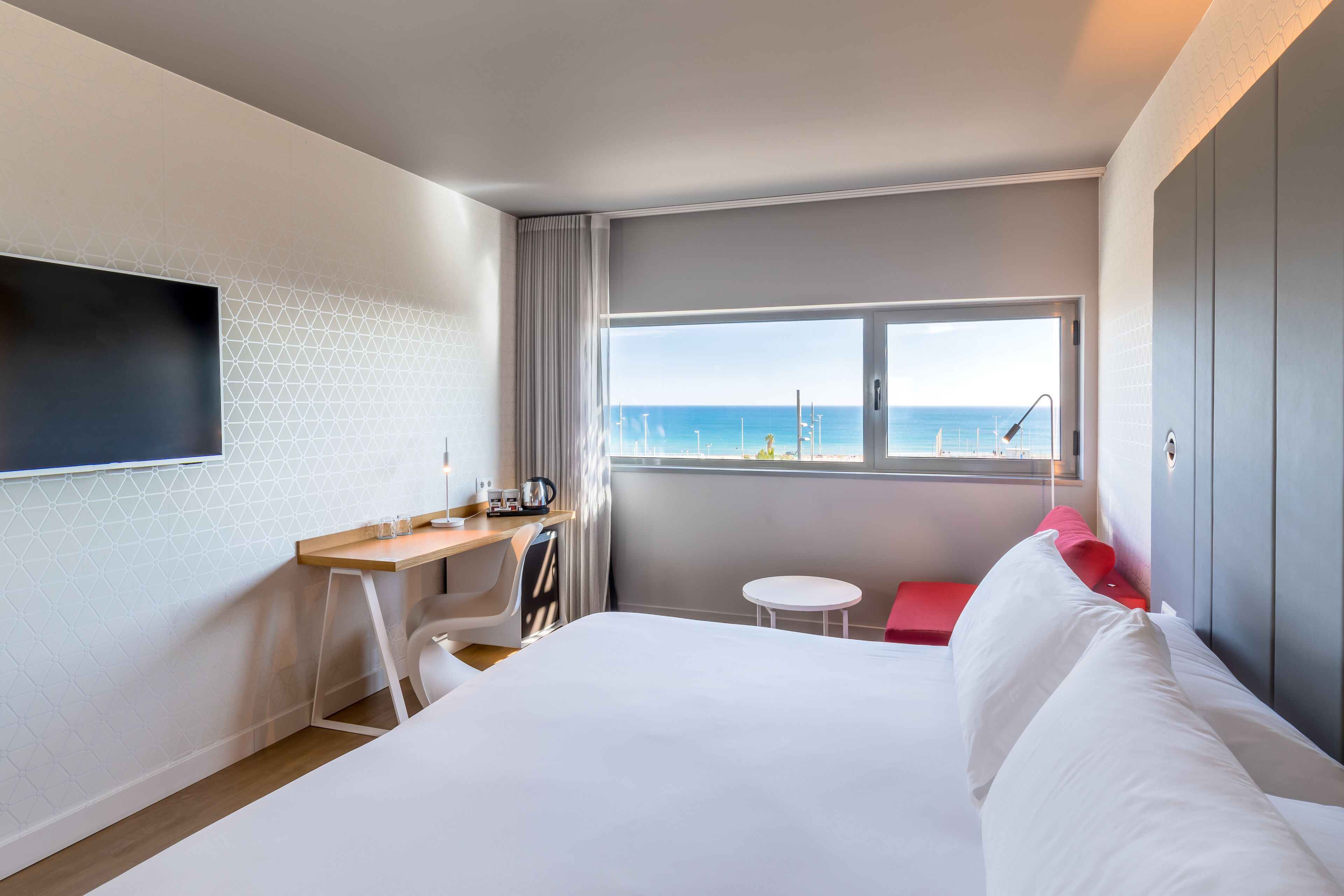 Habitación de hotel con cama, escritorio, silla, una mesita, televisión montada en la pared y ventana con vista al mar.