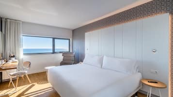 Een moderne hotelkamer met een groot bed, een bureau met stoel, een bijzettafeltje en uitzicht op zee door een groot raam.