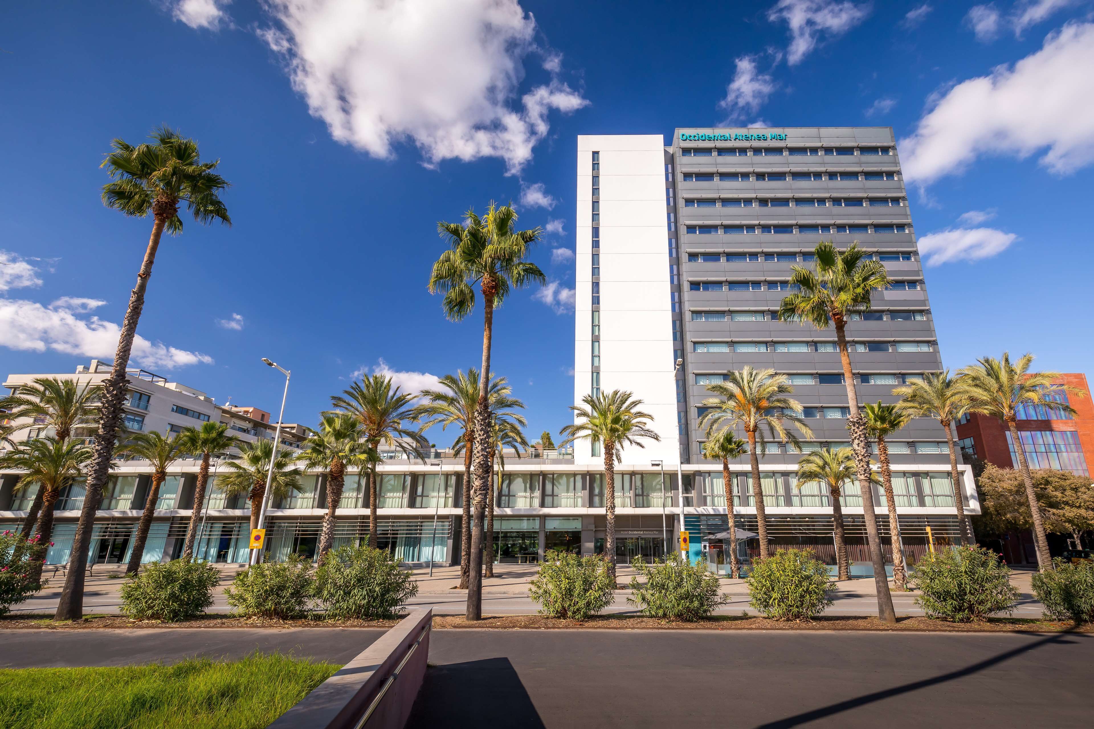 A modern hotel with a prominent vertical tower, surrounded by palm trees and a clear blue sky.