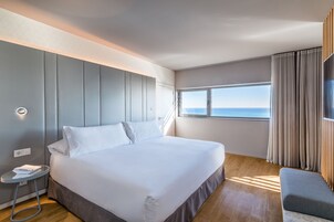Quarto de hotel moderno com uma cama grande, uma mesinha lateral, um espaço de estar e vista para o mar.