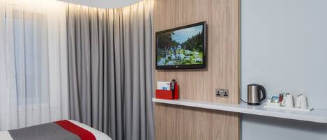 Une chambre d’hôtel avec deux lits, un téléviseur fixé au mur, un panneau en bois, une étagère avec une bouilloire et des tasses, et un rideau.