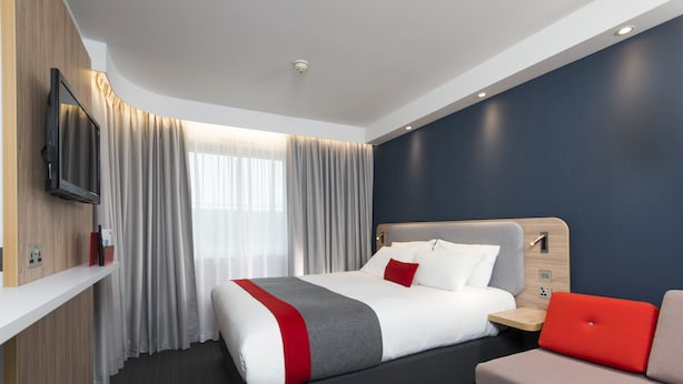 A modern hotel room with a large bed, a flat-screen TV mounted on the wall, a bedside table, and a red upholstered chair. - Holiday Inn Express Cambridge by IHG (Cambridge)