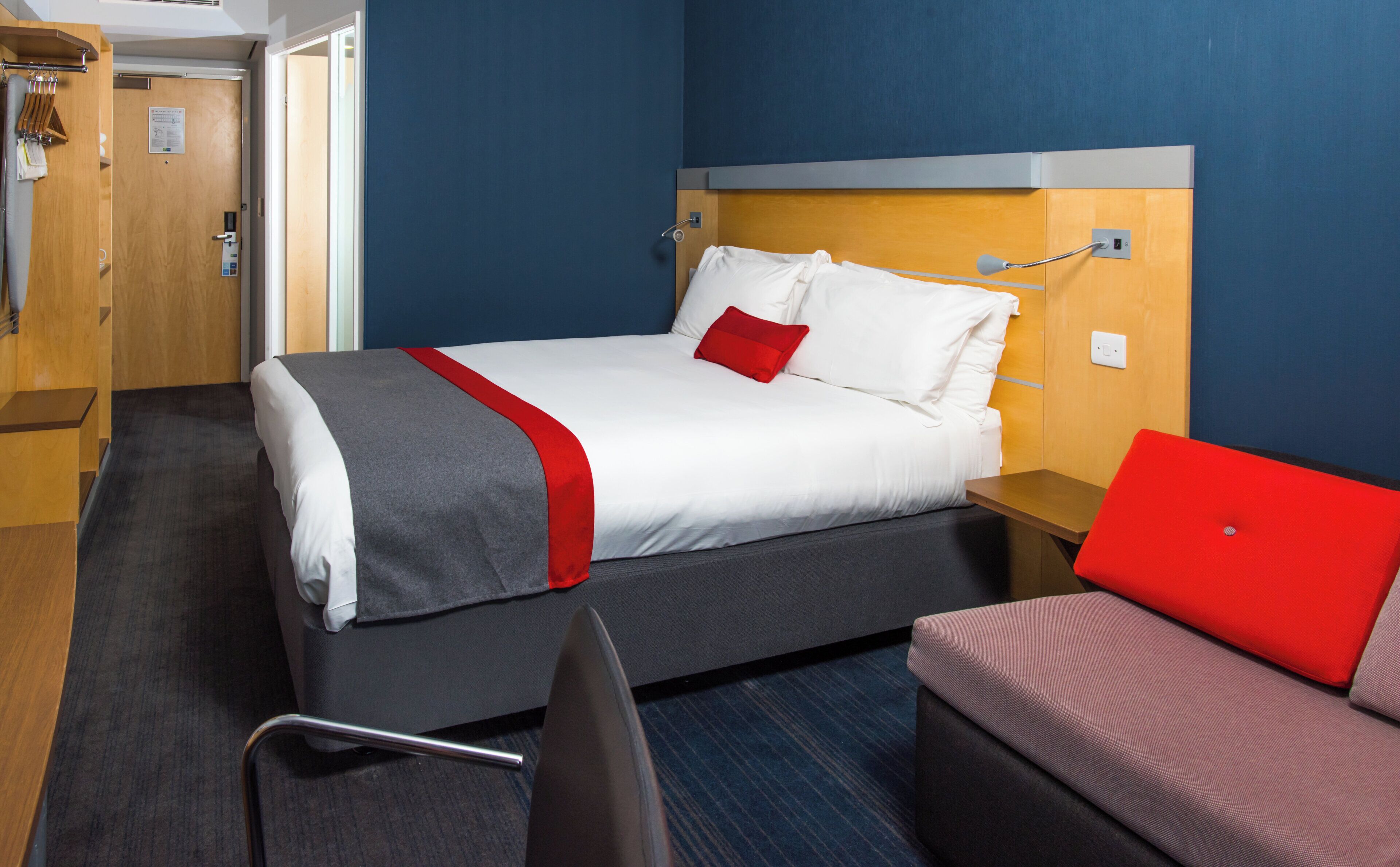 A hotel room with a bed, a red chair, a wooden wardrobe, and a bathroom door.