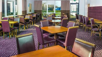 Un comedor de hotel con varias mesas y sillas, ventanas amplias con cortinas y vistas al exterior.