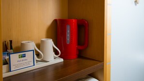 A hotel room with a wooden shelf holding a red kettle, a white mug, and a sign for hot drinks. There are towels stacked below the shelf. - Holiday Inn Express Cambridge by IHG (Cambridge)