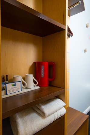 A hotel room with a wooden shelf holding a red kettle, a white mug, and a sign for hot drinks. There are towels stacked below the shelf. - Holiday Inn Express Cambridge by IHG (Cambridge)