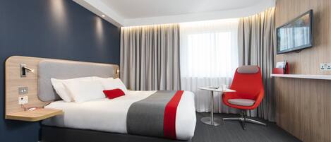 A modern hotel room with a bed, a desk, a chair, and a TV on the wall.