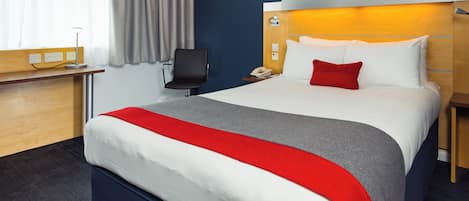 A hotel room with a bed, a desk, a chair, and a nightstand with a lamp.