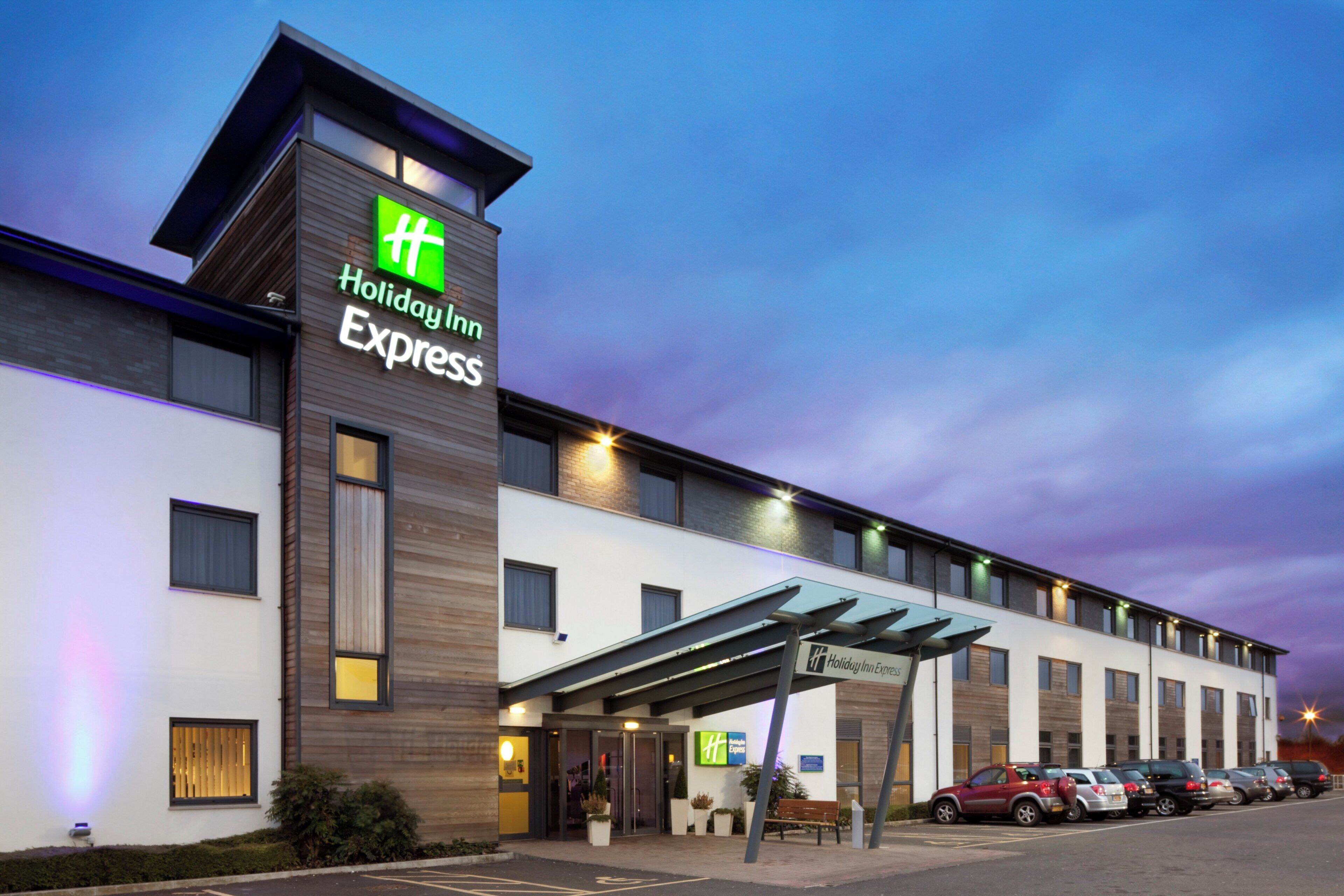 Un edificio de Holiday Inn Express con un diseño moderno, que presenta un logotipo verde llamativo, una entrada cubierta y un estacionamiento con varios autos.