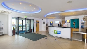A hotel reception area with a modern design, including a reception desk, a waiting area, and a glass door entrance. - Holiday Inn Express Cambridge by IHG (Cambridge)