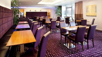 A hotel dining area with multiple tables and chairs, a patterned carpet, and a large window with curtains.