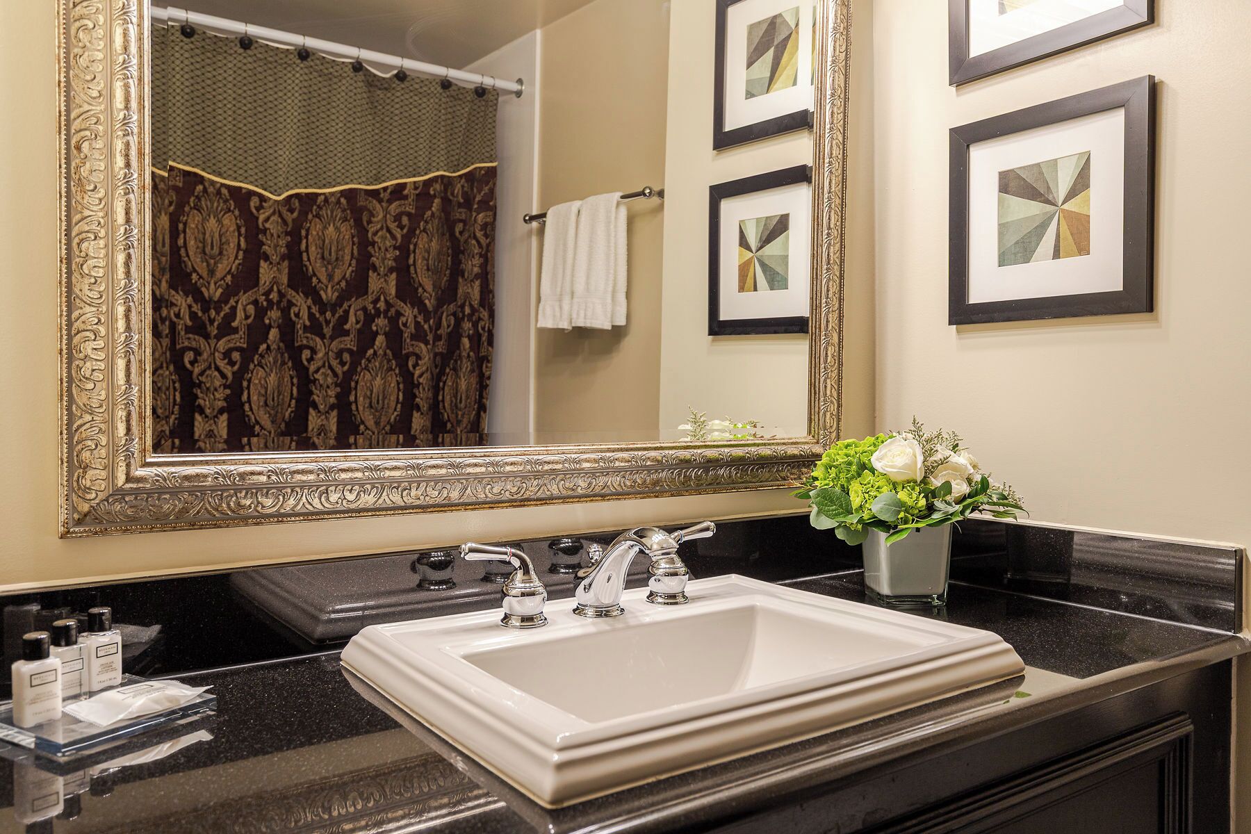 A bathroom with a large ornate mirror, a white sink, and a vase with white flowers.