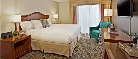 A hotel room with a large bed, a desk with a flat-screen TV, a chair, and a small table with a lamp.