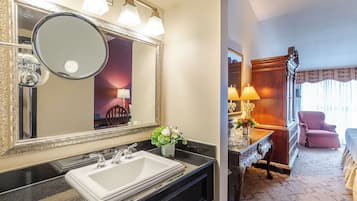 A hotel bathroom with a large mirror, a sink, and a vanity. There is a wooden chair and a lamp in the adjacent room.