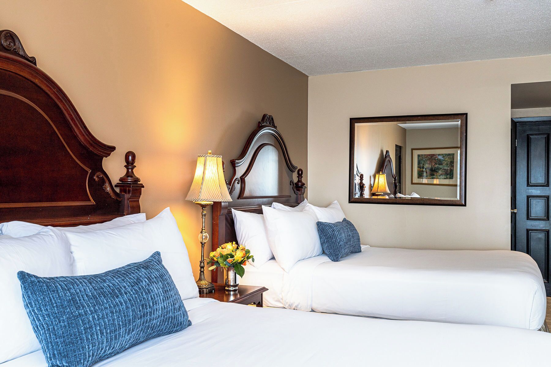 Une chambre d’hôtel avec deux lits, une tête de lit en bois, une table de chevet avec une lampe, un vase de fleurs et un miroir au mur.