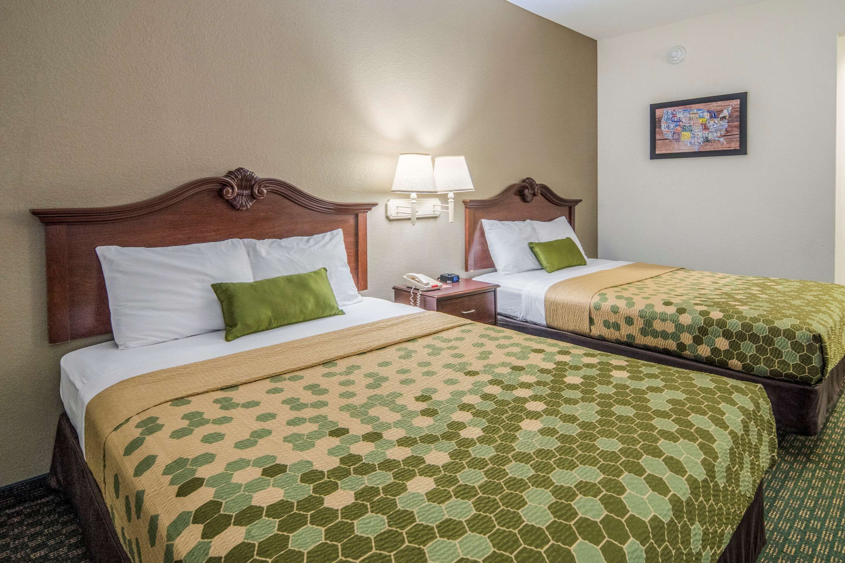 A hotel room with two beds, each with a green and beige patterned bedspread, a wooden headboard, a nightstand with a lamp, and a framed map on the wall.
