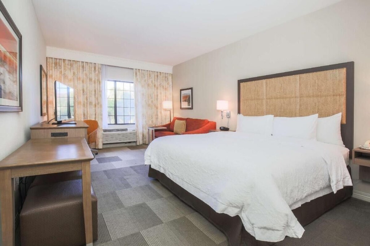 A hotel room with a large bed, a desk, a chair, a small table, and a window with curtains.