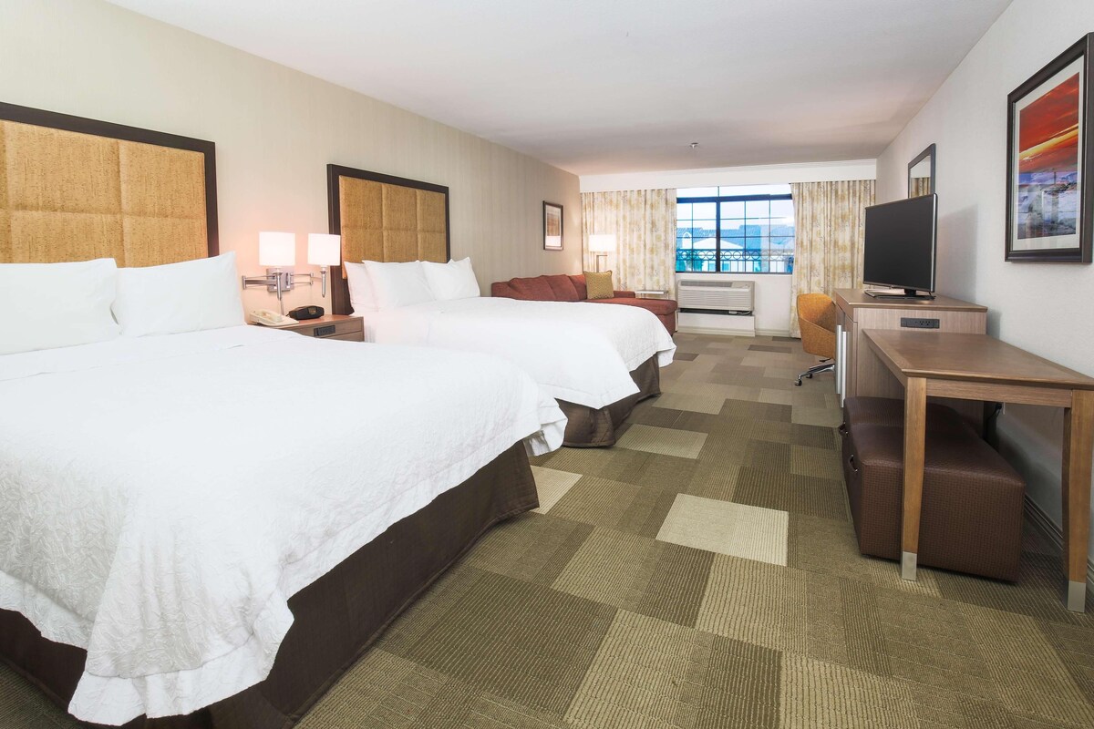 A hotel room with two beds, a desk, a television, and a window with curtains.