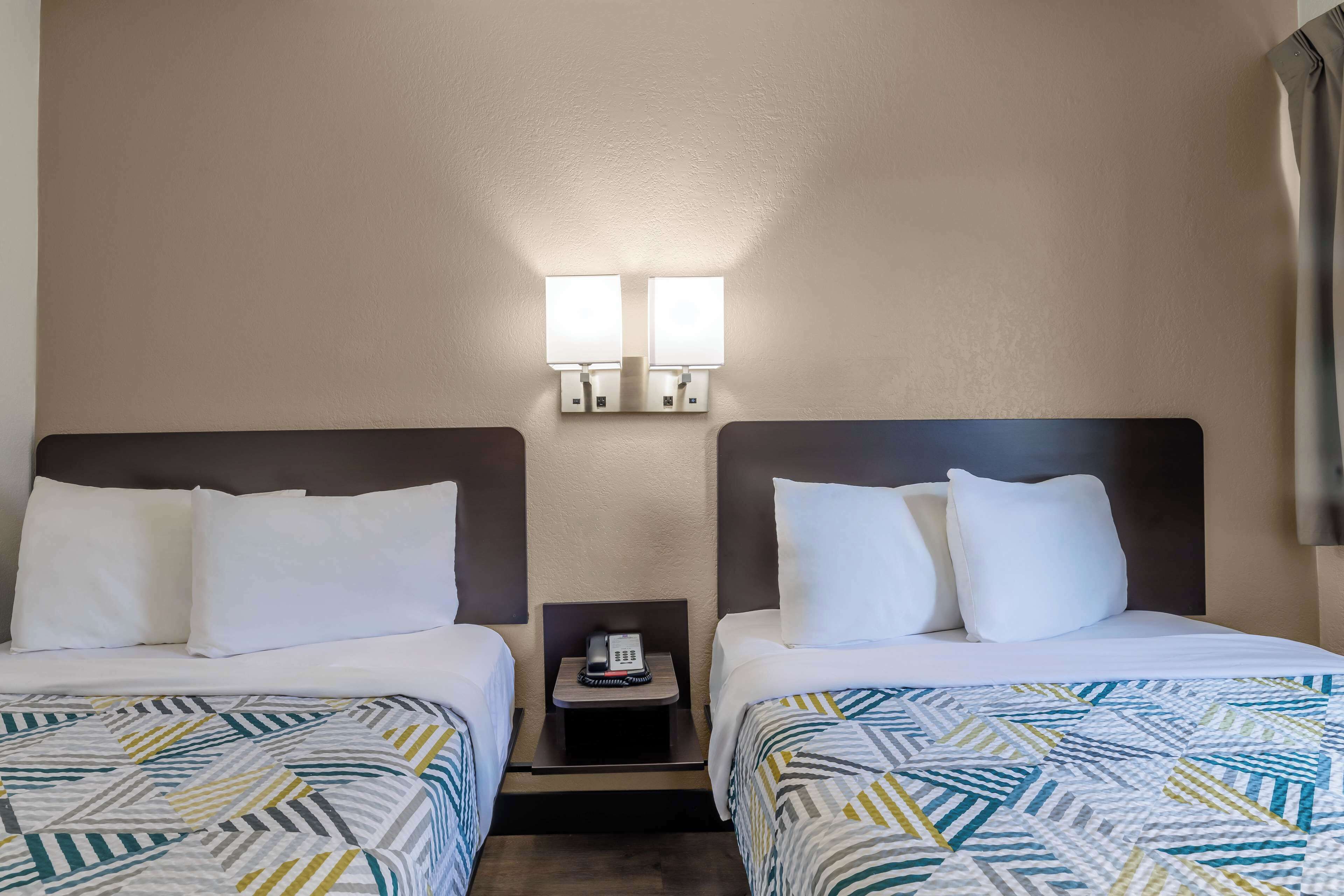 Une chambre d’hôtel avec deux lits, chacun étant équipé de draps blancs et d’une couette à motifs. Il y a deux tables de chevet, chacune avec une lampe, et un téléphone est posé sur l’une d’elles. Les murs sont de couleur beige, et un rideau se trouve sur le côté droit.