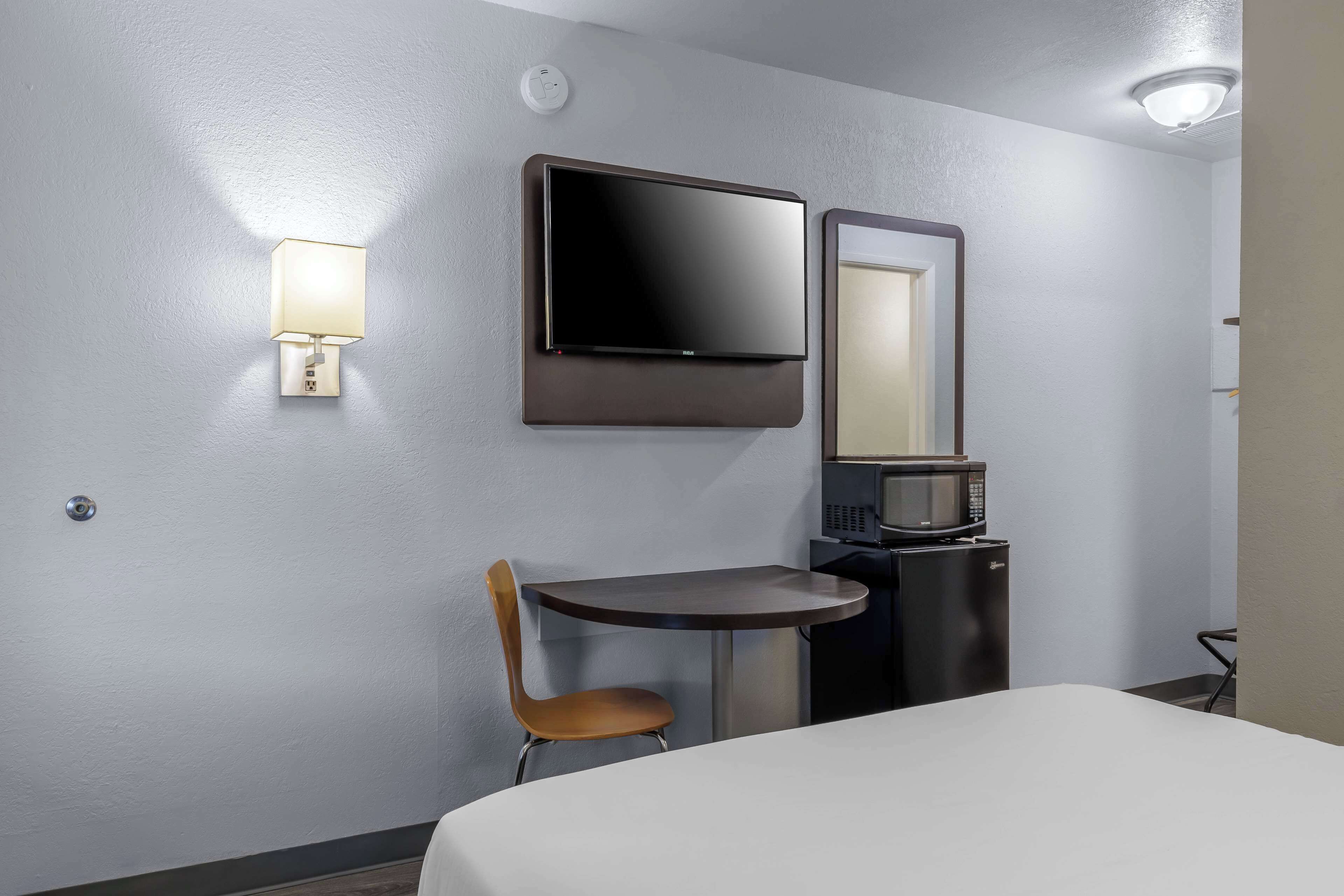 Quarto de hotel com cama, escrivaninha com cadeira, televisão de tela plana na parede, micro-ondas e uma pequena mesa.
