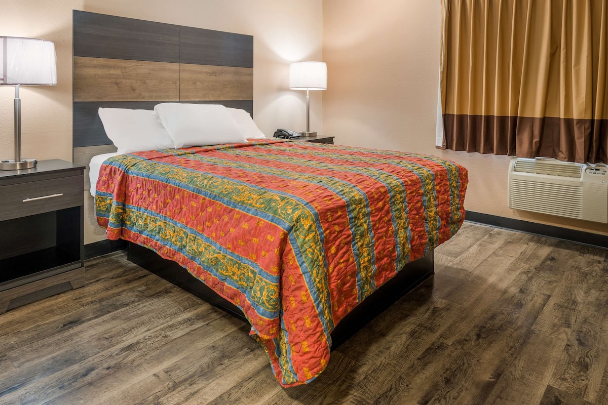A hotel room with a wooden floor, a bed with a colorful quilt, a nightstand with a lamp, and a window with curtains.