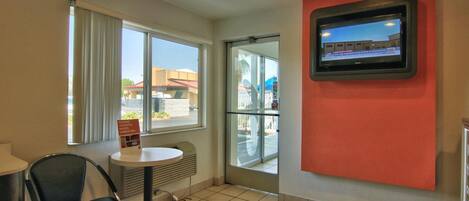 A hotel room with a television mounted on the wall, a small table with a chair, and a window with curtains.