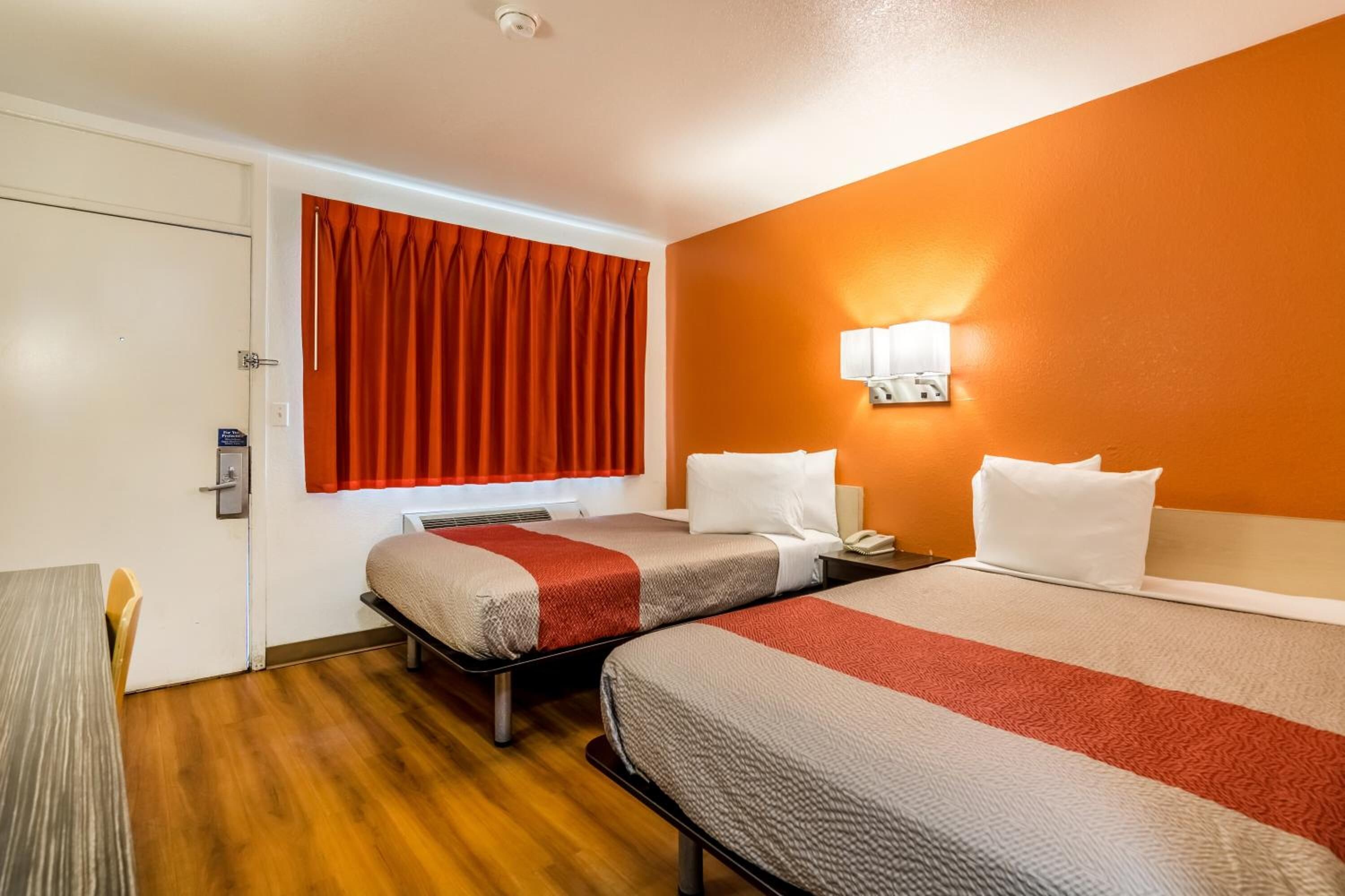 A hotel room with two beds, a wooden floor, a window with red curtains, and a wall-mounted lamp.