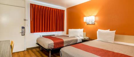 A hotel room with two beds, a wooden floor, a window with red curtains, and a wall-mounted lamp.