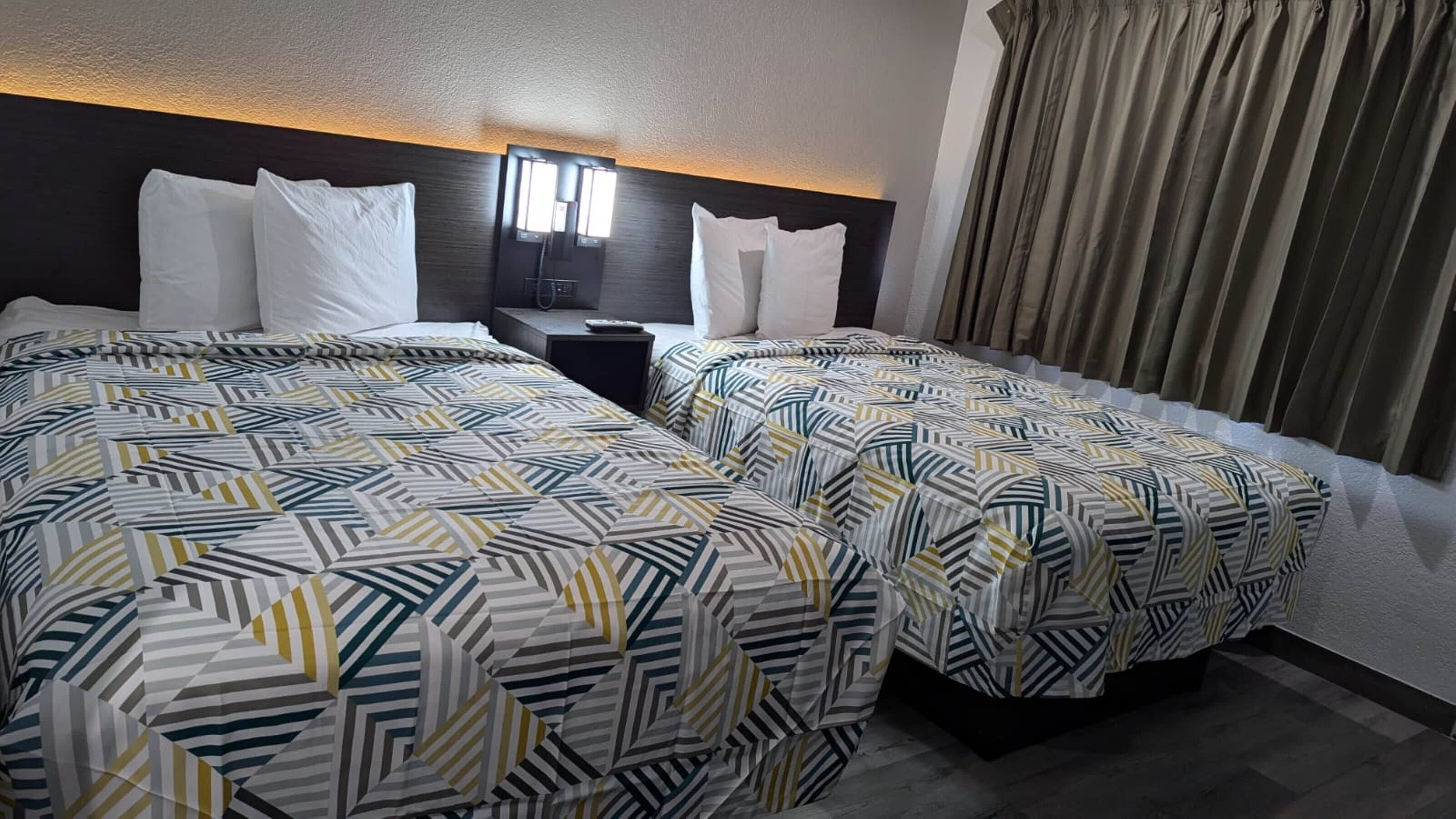 A hotel room with two beds, each with a patterned bedspread, a bedside table with a lamp, and a window with curtains.