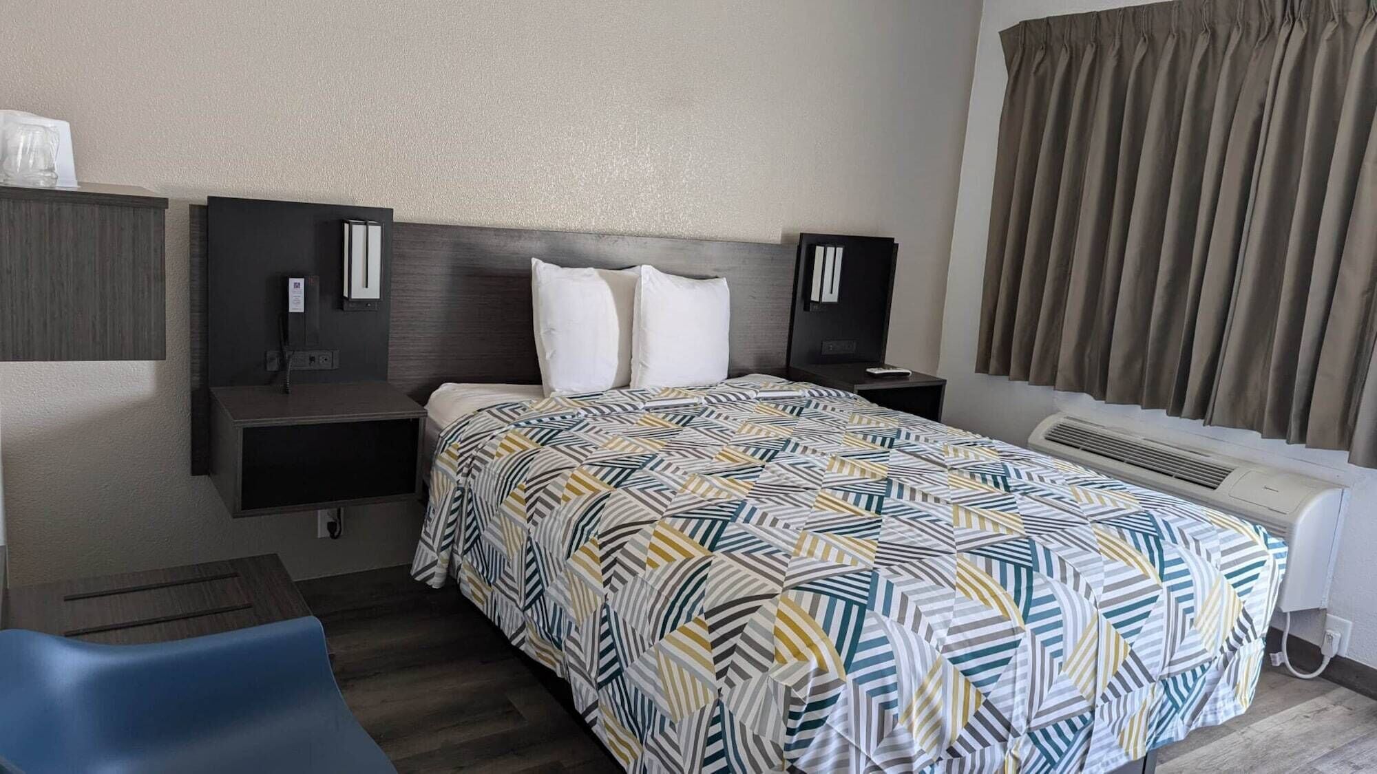 A hotel room with a bed, two bedside tables, a chair, a desk, a window with curtains, and an air conditioning unit.