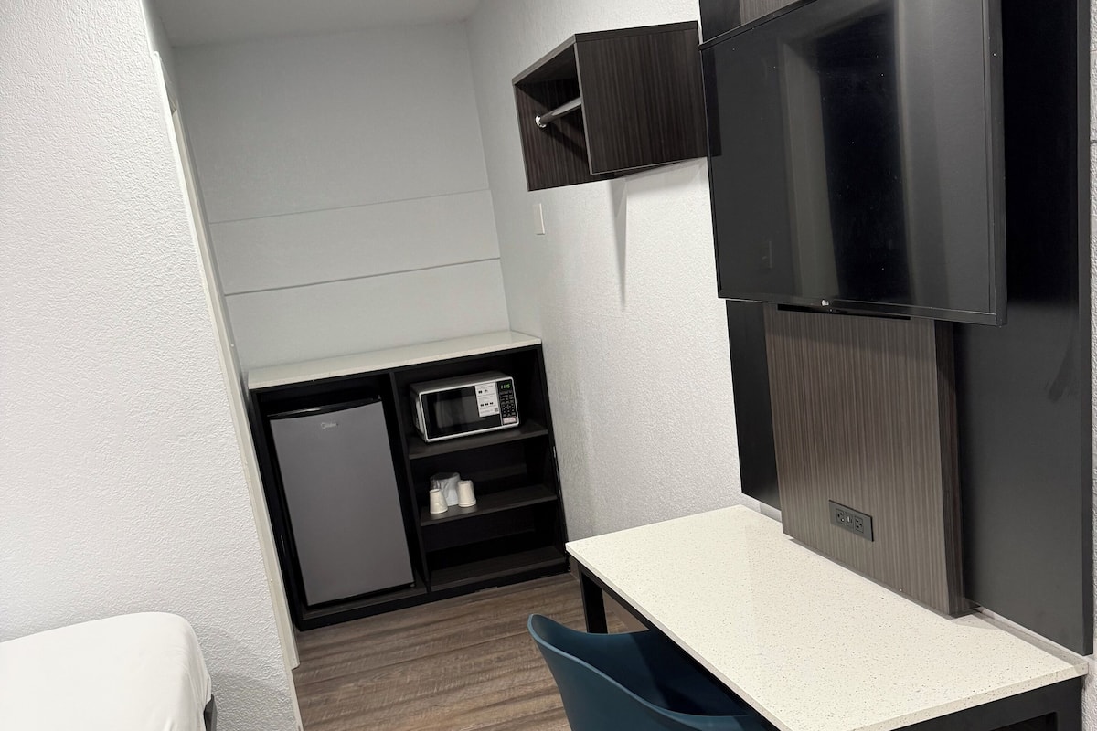 A compact hotel room with a bed, a desk with a chair, a TV mounted on the wall, a microwave, and a small refrigerator.