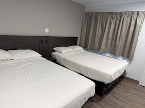 Hotellihuone, jossa on kaksi sänkyä, sängynpääty, seinään asennettu ilmastointilaite ja ikkuna, jossa on verhot.